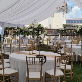 Outdoor event setup with draped white tent, floral centerpieces, and gold chiavari chairs.